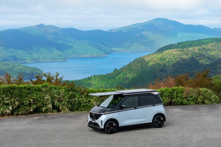 Nissan Sakura electric vehicle is parked on a gray paved road with lush green mountains in the background. The car faces left and features a two-tone body in white and black. Its roof-mounted solar panel is extended forward. Dense greenery and shrubs fill the backdrop, highlighting the car in a natural setting.