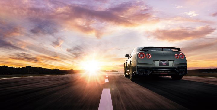 A 2024 Nissan GT‑R driving on a straight road towards a sunset with trees on the left hand side.