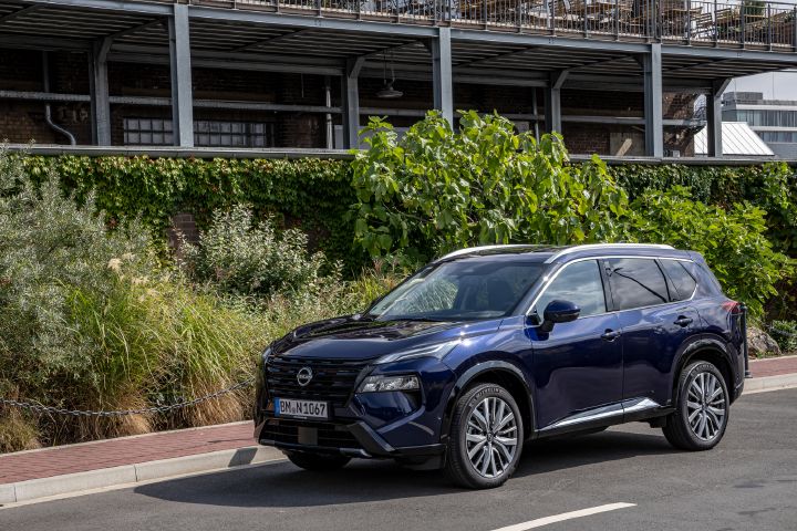 Statische 3/4 Frontaufnahme eines dunkelblauen Nissan X‑Trail, der vor einer begrünten Mauer hält