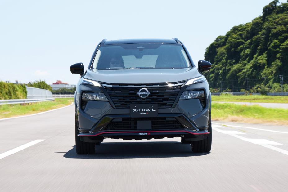 A grey Nissan X-Trail NISMO is driving on a road. The vehicle is captured from the front, with greenery and trees in the background.