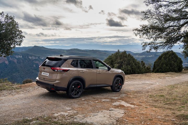 Neuer 2026 Nissan X‑Trail in der Karosseriefarbe „Baja Storm”, geparkt auf einem Feldweg mit Blick auf eine bergige Landschaft und bewölkten Himmel. Das Fahrzeug verfügt über schwarze Felgen und ein schwarzes Dach und steht in der Nähe von Bäumen mit weitläufigem Tal und Hügeln im Hintergrund.