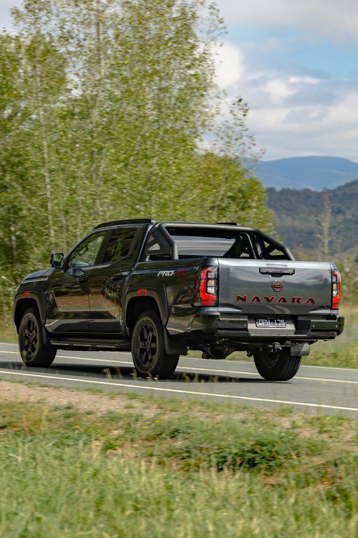 2026 Nissan Navara PRO-4X finished in Boulder Grey featuring a portrait rear three-quarter scene with black sports bar and red NAVARA tailgate lettering.