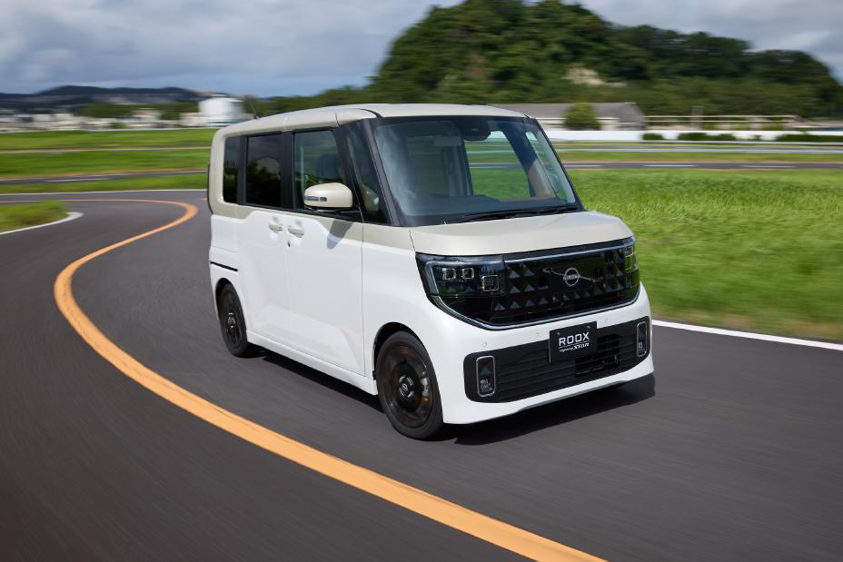 The new Nissan Roox kei car, featuring a white pearl / frozen vanilla pearl two-tone, is driving on a curved road. The background includes green grass, trees, and buildings in the distance under a partly cloudy sky.