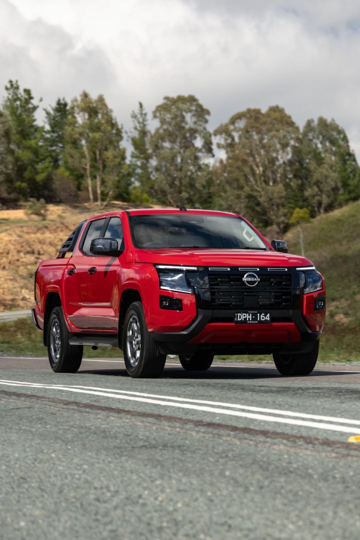 Front view of a 2026 Nissan Navara ST‑X dual cab in Outback Red, travelling along a sealed rural road lined with trees and open fields.