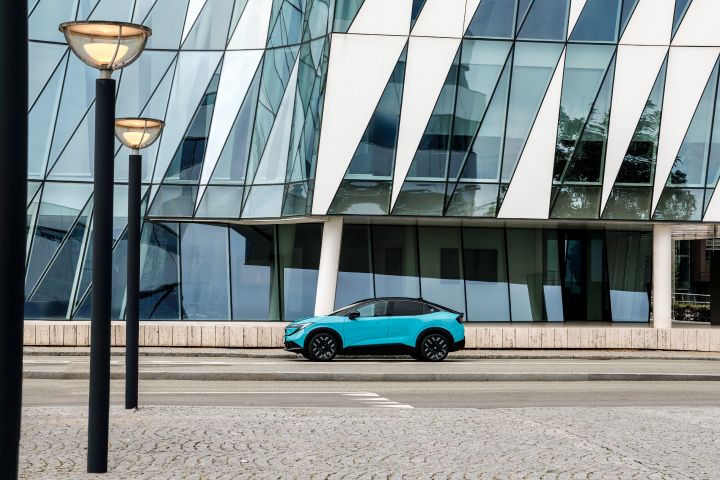 Klein in der Mitte des Bildes die Seitenansicht eines türkisfarbenen Nissan LEAF, im Hintergrund eine moderne Glasfassade und im Vordergrund ein gepflasterter Platz