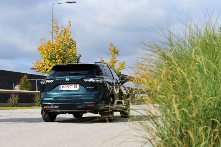 3/4 Heckansicht eines dunkelgrünen Nissan Qashqai e‑Power, der bei Sonnenschein auf einem Parkplatz steht, die rechte Bildhälfte ist von hohen grünen Ziergräsern ausgefüllt