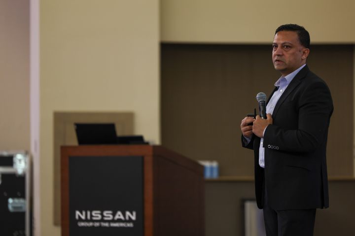 A man in a black suit speaking into a microphone with his hands near his chest.