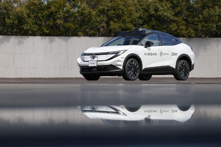 A white Nissan LEAF is parked outdoors at a front‑diagonal angle, with its reflection visible on the pavement in the foreground. Black “NISSAN,” “Wayve,” and “Uber” logos appear on the side. A light gray concrete wall and green trees form the background under a calm sky, highlighting the vehicle’s white exterior.