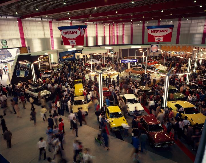wide angle shot of a bustling car exhibition with various vehicles on display and large Nissan banners handing from the ceiling.