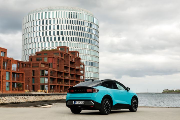 3/4 Heckansicht eines türkisen Nissan LEAF mit schwarz abgesetztem Heckfenster, das Auto steht an einem Kai, im Hintergrund sieht man markante braune und gläserne Gebäude