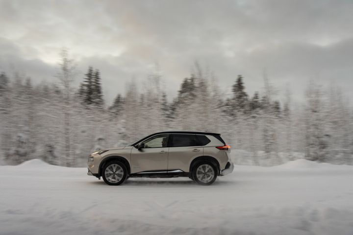 Silberner Nissan X‑Trail e‑Power e‑4ORcE fährt auf schneebedeckter Straße mit verschneiten Bäumen im Hintergrund, Seitenansicht