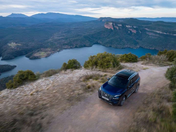 Der neue Nissan X‑Trail SUV 2026 in der Außenfarbe Universal Blue fährt auf einer unbefestigten Straße entlang eines Hügels mit Blick auf einen großen See, der von bewaldeten Bergen umgeben ist. Die Szene unterstreicht das Outdoor-Abenteuer und die Leistungsfähigkeit des Fahrzeugs mit natürlichen Farben wie Grün, Wasser und entfernten blauen Bergketten unter einem bewölkten Himmel.
