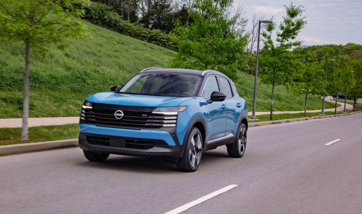Front left 3/4 view of a blue 2025 Nissan Kicks driving along a road with vibrant grass and trees alongside.