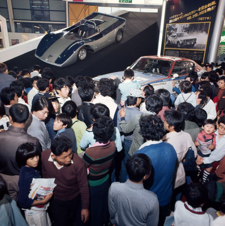 A vintage open-top Nissan car with only one driver seat. It is surrounded by a crowd of onlookers at the auto show.