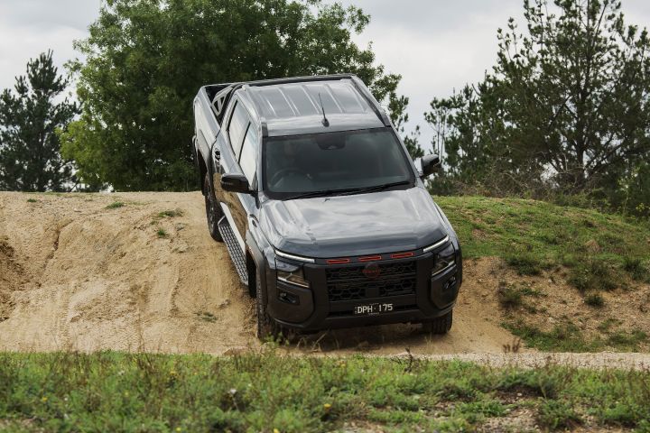 2026 Nissan Navara PRO-4X finished in Boulder Grey pictured in a three-quarter downhill view dropping off a sandy ledge with roof rails, sports bar and carved wheel ruts.