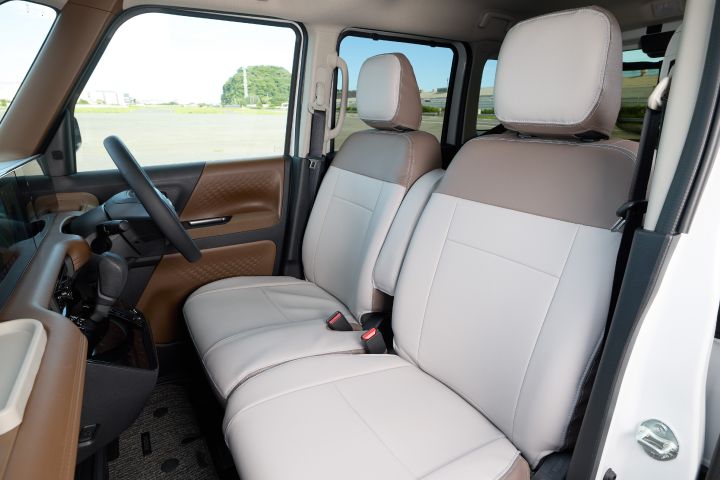 Interior view of the new Nissan Roox kei car. The front seat headrests feature a greige and brown two-tone design, with outdoor scenery visible through the car windows in the background.