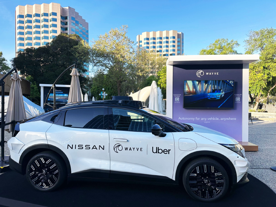 Ein weißer Robotaxi-Prototyp auf Basis des neuen Nissan Leaf ist in einem Ausstellungsbereich im Freien von der Seite zu sehen. Im Hintergrund sind Bäume, hohe Gebäude und ein Stand mit einem großen Bildschirm zu erkennen.