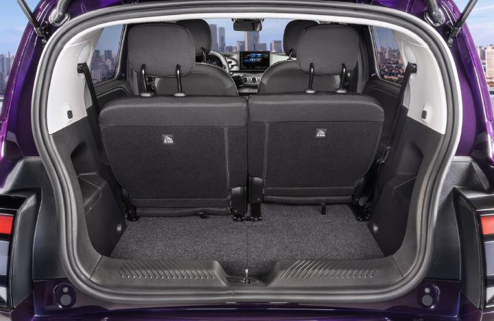 Fotografi som viser kompakt hatchback-bagasjerom med oppreist bakseter, svart trekk, teppebelagt gulv og vidåpen bakluke. Lilla eksteriør, delte seterygger, bakre nakkestøtter, dashbord og byens silhuett synlig, og fremhever tilgjengelig lagringsplass og bilinteriør.