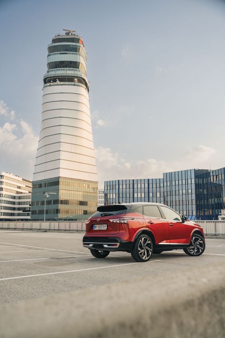 Blick von schräg hinten auf einen Nissan Qashqai in Fuji Sunset Red vor einem hohen, weißen Gebäude