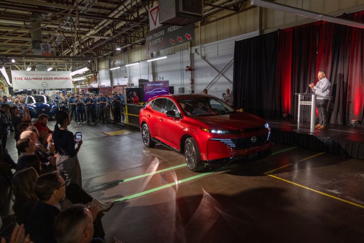 A red 2025 Nissan Murano parked between a stage and a crowd of workers, a spotlight shines directly on it.
