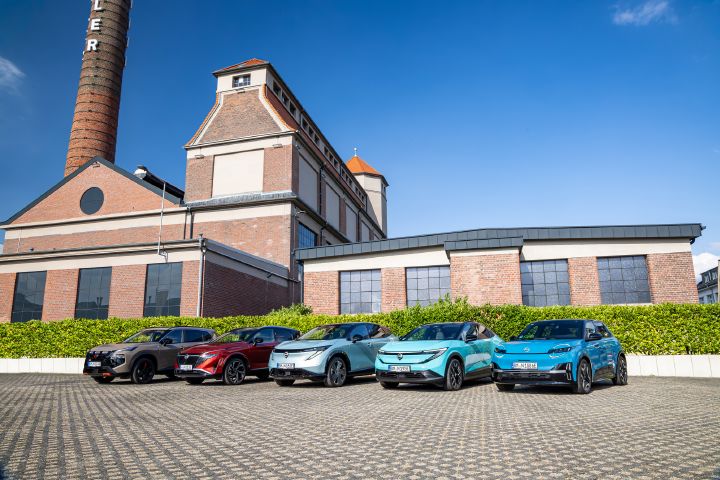 Vor einem Backsteingebäude und einer grünen Hecke stehen nebeneinander die fünf Nissan Modelle Nissan X‑Trail N-Trek, Nissan Qashqai, Nissan Ariya, Nissan Leaf und Nissan Micra, 3/4 Frontansicht