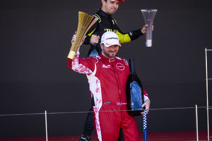 Oliver Rowland in seinem roten Rennanzug des Nissan Formula E Teams, er hält in der rechten Hand den Siegerpokal des Monaco E-Prix hoch und in der linken Hand eine Champagnerflasche.