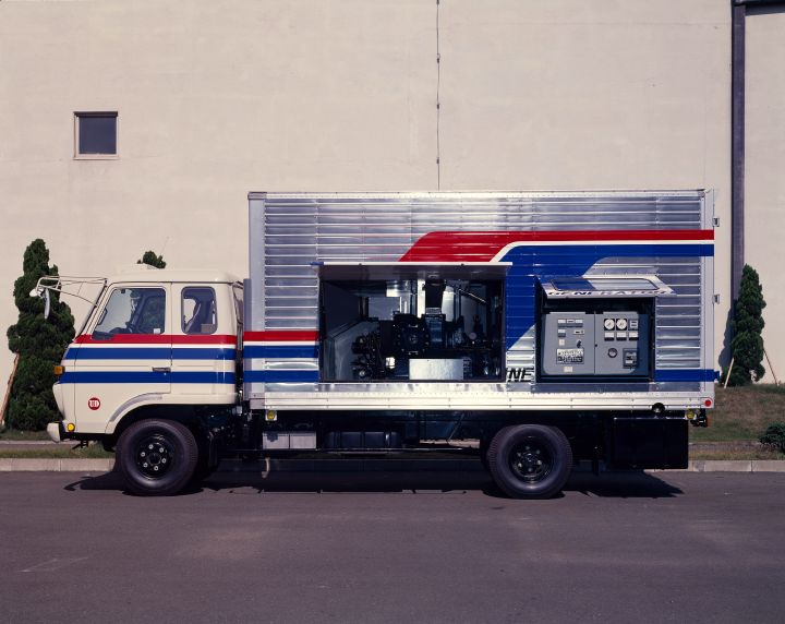 a gas turbine truck parked outside of a large building with a prominent red and blue stripe running across the side of the truck and large black letters with the text Nissan Gas Turbine Generator written across it. The flaps are open showing mechanical parts of the generator inside.