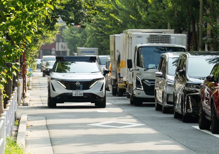 Ein weißer Nissan Ariya fährt auf der linken Straßenseite vorwärts, die Straße ist von Bäumen gesäumt und am Rand parken Fahrzeuge. Im Hintergrund ist die Skyline von Tokio zu sehen.