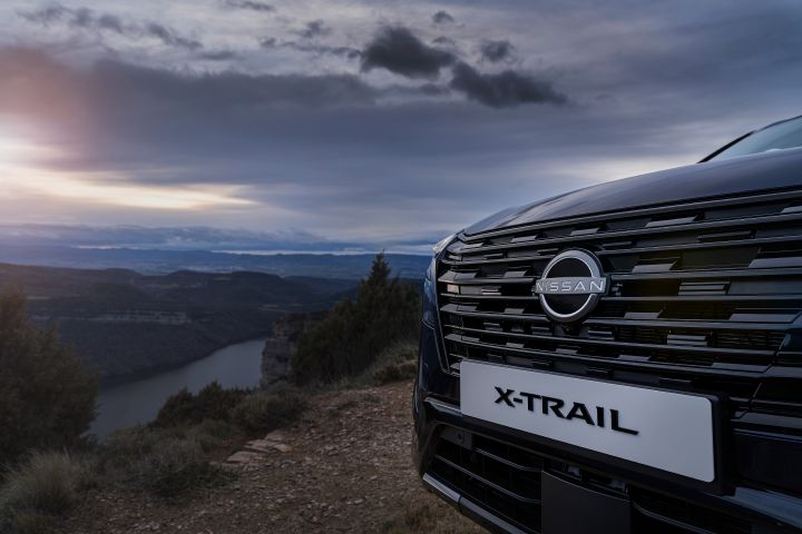 Neuer 2026 Nissan X‑Trail Tekna in der universellen Außenfarbe Blau mit Nahaufnahme des Kühlergrills und Emblems eines schwarzen Nissan X‑Trail SUV, der bei Sonnenuntergang auf einer Klippe mit Blick auf ein Flusstal geparkt ist. Die Szene unterstreicht die raue Outdoor-Umgebung mit dramatischem Himmel und fernen Bergen und betont das abenteuerliche und robuste Design des Fahrzeugs.