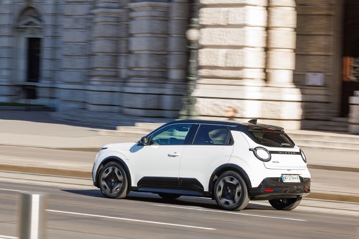 7/8 Heckansicht eines weißen Nissan Micra mit schwarzem Kontrastdach, der zügig eine Straße entlang fährt mit historischen Gebäuden im Hintergrund
