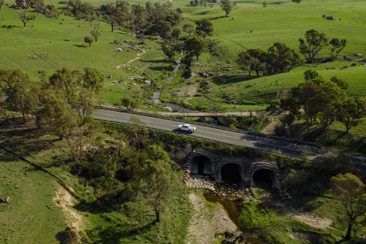 2026 Nissan Navara SL dual cab ute finished in Alpine White seen from above crossing a bridge over stone culverts and a narrow rural creek.