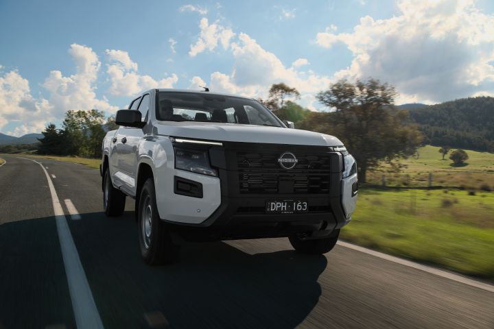 2026 Nissan Navara SL dual cab ute finished in Alpine White captured in motion from the front three-quarter angle on a country road with hills and open paddocks.