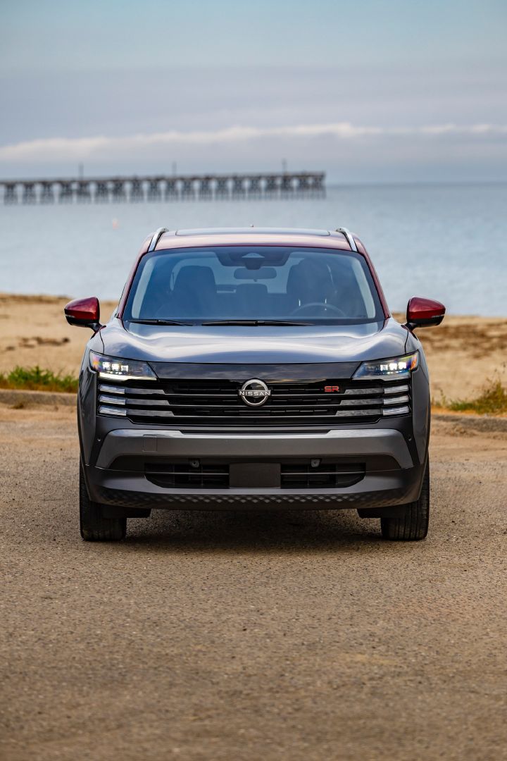 Front view of a silver and red accented 2025 Nissan Kicks parked on a gravel lot overlooking a beach, ocean and Pier.
