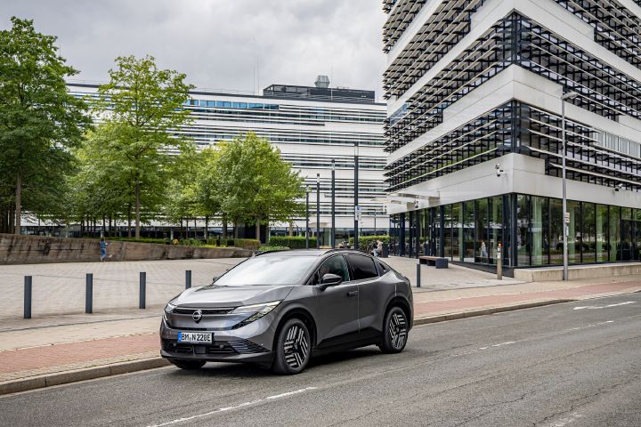 Vor mehreren modernen, städtischen Hochhäusern steht ein grauer Nissan Leaf, 3/4 Frontansicht
