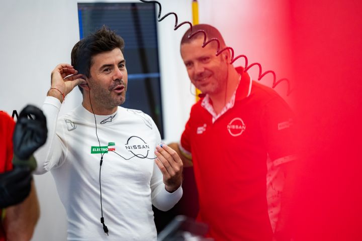 Norman Nato steht in einem weißen Rennanzug da und richtet gerade seinen Ohrhörer zurecht. Rechts ist eine weitere Person in einem roten Nissan Team-Shirt zu sehen, im Hintergrund ist eine Boxengasse in einer Halle zu erkennen. Ein rotes, unscharfes Element im Vordergrund rahmt die Szene teilweise ein.