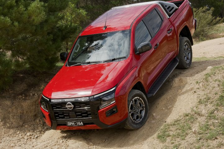 A 2026 Nissan Navara ST‑X dual cab finished in Outback Red navigating an uneven sandy off‑road hill with pine trees in the background.