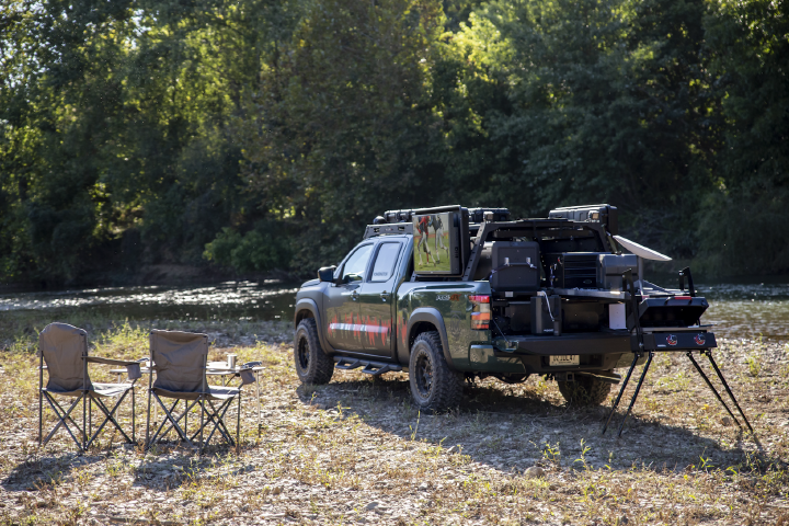 A green 2025 Project Trailgater Nissan Frontier parked on a tree-lined grass field with two camping chairs.