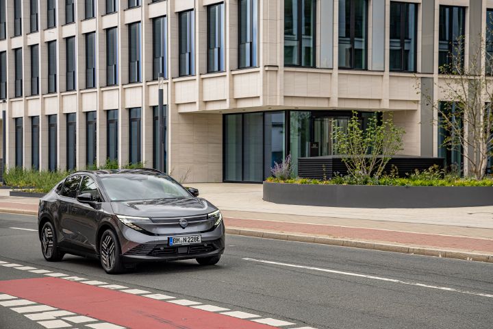 Vor einem Gebäude mit heller Front und vielen Fenstern steht schräg ein grauer Nissan Leaf, durch den Bildvordergrund verläuft ein rot abgesetzter Radweg