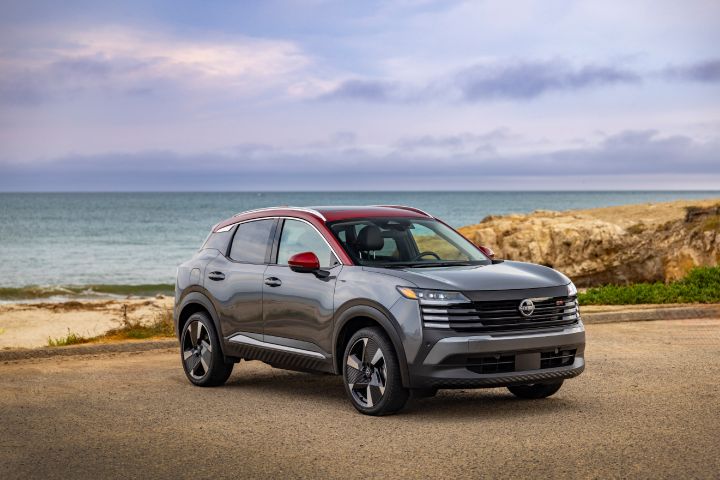 Front right 3/4 of a silver 2025 Nissan Kicks parked on a gravel lot overlooking sand and ocean on a cloudy day.