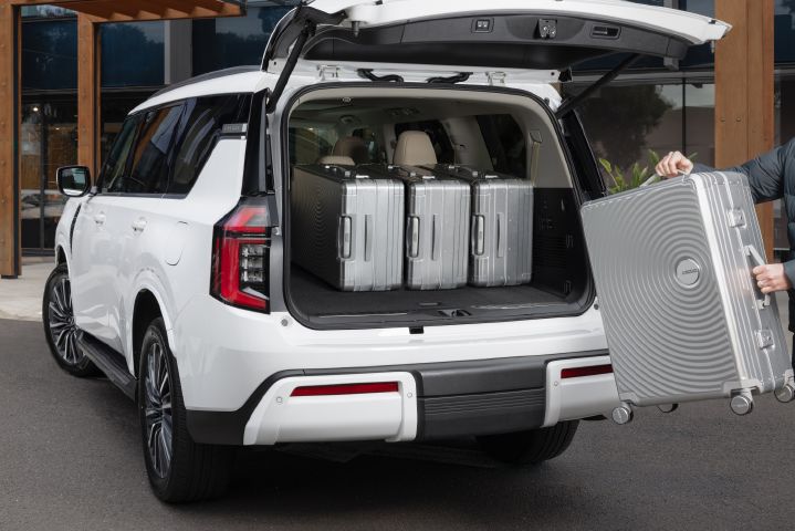 2026 Nissan Y63 Patrol finished in white, shown from a rear three-quarter exterior angle with the tailgate open, four large silver suitcases loaded in the cargo area and another suitcase being loaded.