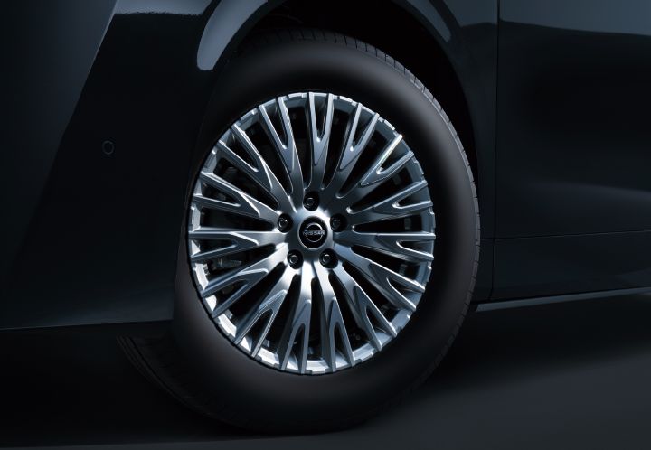 ブラックのエルグランド VIPのホイール周辺を、車両側面からクローズアップで撮影した画像。画面中央には大径のシルバーカラーのアルミホイールが配置されており、多数の細いスポークが放射状に広がる精緻で高級感のあるデザインとなっている。ホイール中央には日産エンブレムが配置され、スポークの間からブレーキディスクやブレーキキャリパーの一部が見える。
ホイールを囲むタイヤはブラックで、トレッドやサイドウォールの質感が確認できる。周囲には光沢のあるブラックボディが写り込み、上部にはホイールアーチ、右側にはサイドパネルが見えている。車両の向きは画面右方向で、側面下部のボディラインやフェンダー形状が確認できる。
ボディカラーは深みのあるブラックで、スタジオ照明を受けた滑らかな反射や陰影によって立体感が強調されている。