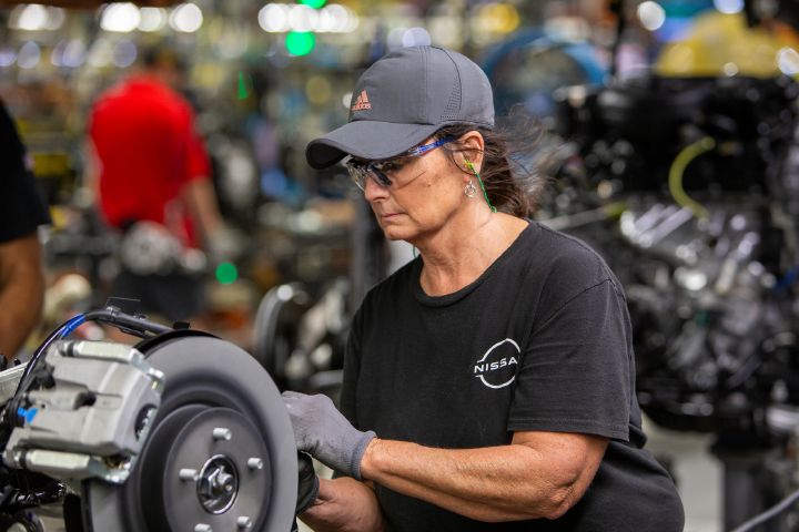 A woman, wearing a gray hat and black Nissan shirt, working on a machine on a manufacturing plant floor.