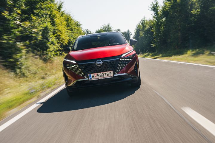 Frontale, dynamische Fahraufnahme eines Nissan Qashqai in Fuji Sunset Red auf einer sonnigen Landstraße mit dichtem Grün an den Fahrbahnrändern