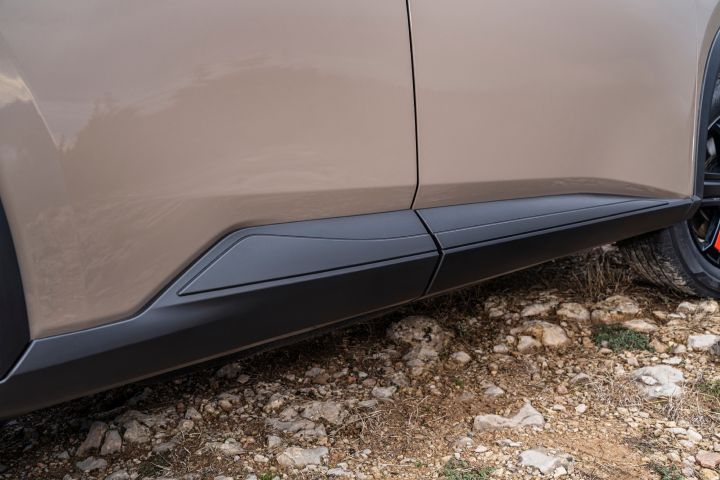 Der neue 2026 Nissan X‑Trail N-Trek in der Karosseriefarbe „Baja Storm” mit Sicht auf die untere Seitenverkleidung und die schwarzen Seitenschweller über felsigem, unebenem Gelände. Das Bild hebt den Kontrast zwischen den glatten Oberflächen des Autos und dem rauen Gelände hervor und betont die Designdetails und die Bodenfreiheit des Fahrzeugs.