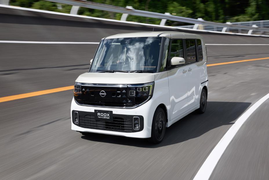 The new Nissan Roox kei car, featuring a white pearl / frozen vanilla pearl two-tone, is driving forward. The background shows a curved highway and guardrails.