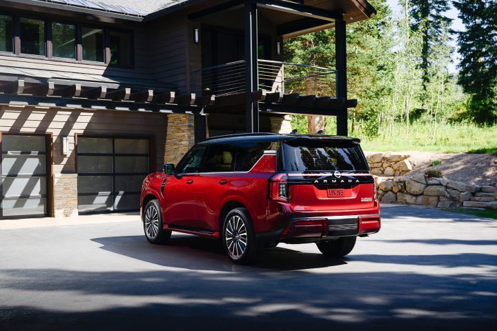 Left rear view of a red 2025 Nissan Armada parked in the driveway of a brick house, greenery is seen in the distance.