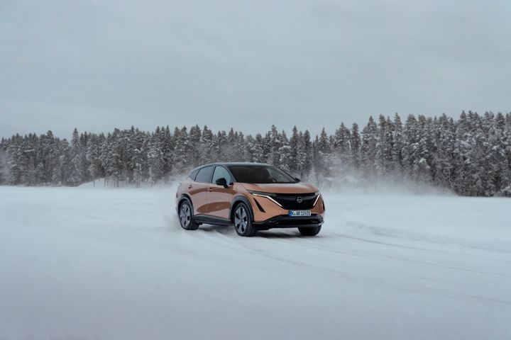 Elektroauto Nissan Ariya e‑4ORCE in Akatsuki Copper mit Dach und Außenspiegel in Pearl Black fährt auf Eissee mit schneebedeckten Bäumen im Hintergrund, 3/4 Front