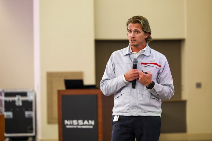 A man in a white windbreaker stand holding a microphone speaking to a crowd in a large room.
