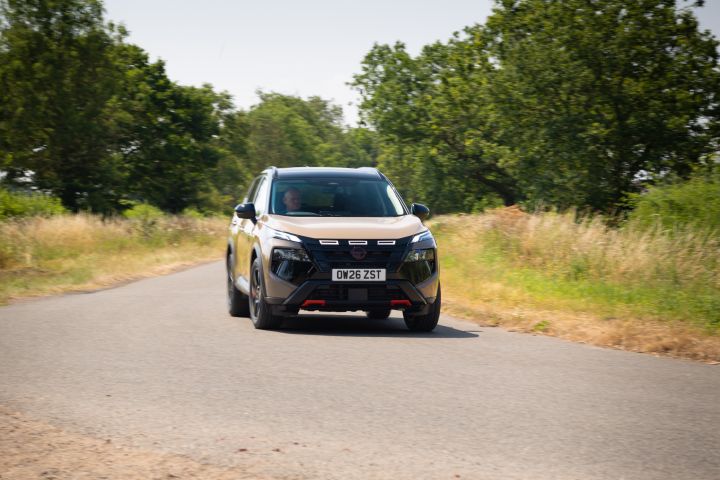 Champagne-coloured Nissan X‑Trail driving along a country road, viewed from the front, with trees and grassland on either side