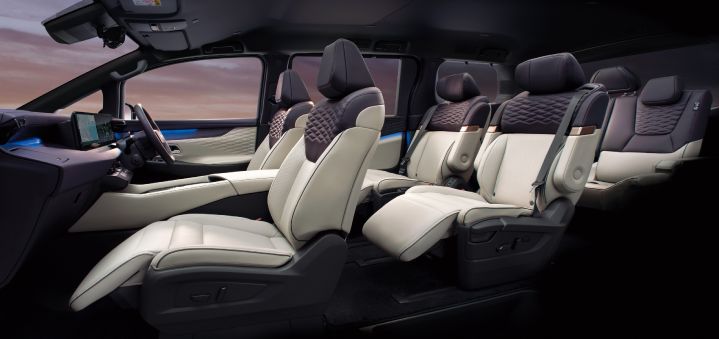 Interior view of the All-new ELGRAND, photographed from the front-left side looking toward the rear of the cabin. The premium interior features a two-tone ivory and dark color scheme, with diamond-pattern upholstery across all three rows of seats. The front area includes a large dashboard display and center console, while the second row is equipped with individual captain’s chairs with the ottomans extended. Blue ambient lighting accents the door panels, and a sunset sky is visible through the side windows. The contrast between the dark headliner and the light-colored seats enhances the spacious and luxurious atmosphere of the cabin.